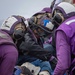 Several personnel wearing purple jerseys and flight deck helmets with goggles are attending to an injured or incapacitated person lying on a wire mesh Stokes litter/stretcher. The scene appears to be a casualty response drill or actual emergency on a naval aircraft carrier flight deck. The responders are wearing standard U.S. Navy aviation boatswain's mate (fuel) purple-colored flight deck gear.
