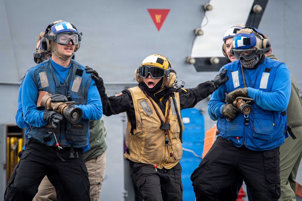 The image shows naval personnel on what appears to be the deck of a military vessel, engaged in what looks like a damage control or firefighting drill. A central figure wearing a yellow helmet and vest appears to be shouting commands or directing others, while personnel in blue jerseys hold firefighting hose nozzles. The scene conveys urgency and coordinated action consistent with carrier deck operations or emergency response training.