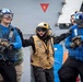 The image shows naval personnel on what appears to be the deck of a military vessel, engaged in what looks like a damage control or firefighting drill. A central figure wearing a yellow helmet and vest appears to be shouting commands or directing others, while personnel in blue jerseys hold firefighting hose nozzles. The scene conveys urgency and coordinated action consistent with carrier deck operations or emergency response training.
