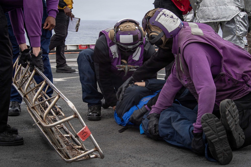 The image depicts what appears to be a military casualty response drill or training exercise on the deck of a naval vessel. Personnel wearing purple jerseys and protective helmets are attending to a simulated casualty lying on the flight deck. A Stokes litter (rescue basket stretcher) is visible nearby, along with observers holding clipboards, suggesting this is a formal training evaluation.