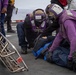 The image depicts what appears to be a military casualty response drill or training exercise on the deck of a naval vessel. Personnel wearing purple jerseys and protective helmets are attending to a simulated casualty lying on the flight deck. A Stokes litter (rescue basket stretcher) is visible nearby, along with observers holding clipboards, suggesting this is a formal training evaluation.