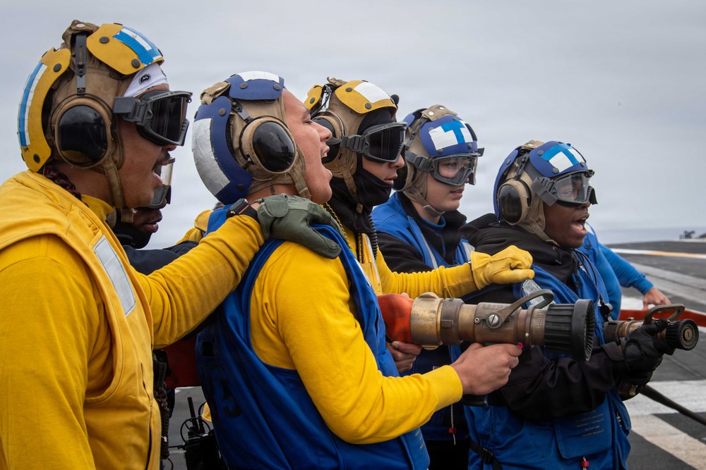 A group of military personnel, likely U.S. Navy sailors, are conducting a firefighting or damage control drill on what appears to be an aircraft carrier flight deck. They are wearing color-coded jerseys and helmets with ear protection and goggles, consistent with carrier deck crew uniforms. Several personnel are holding a large brass fire hose nozzle and appear to be shouting commands or responding to a simulated emergency.