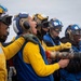 A group of military personnel, likely U.S. Navy sailors, are conducting a firefighting or damage control drill on what appears to be an aircraft carrier flight deck. They are wearing color-coded jerseys and helmets with ear protection and goggles, consistent with carrier deck crew uniforms. Several personnel are holding a large brass fire hose nozzle and appear to be shouting commands or responding to a simulated emergency.