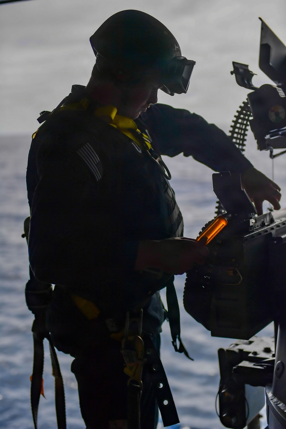 A silhouetted military personnel, identifiable by a U.S. flag patch and yellow safety vest/flotation device, is working with a mounted weapon system, likely a machine gun with an ammunition belt visible. The figure is backlit against what appears to be an open ocean or water background, creating a strong contrast silhouette effect. The operator appears to be loading or performing maintenance on the weapon system.