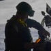 A silhouetted military personnel, identifiable by a U.S. flag patch and yellow safety vest/flotation device, is working with a mounted weapon system, likely a machine gun with an ammunition belt visible. The figure is backlit against what appears to be an open ocean or water background, creating a strong contrast silhouette effect. The operator appears to be loading or performing maintenance on the weapon system.