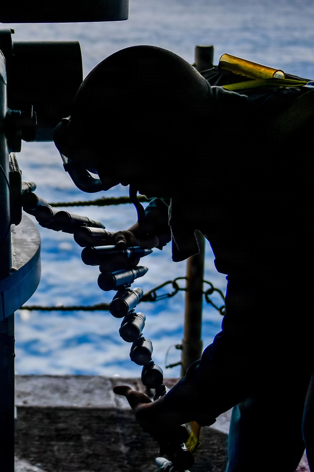 A silhouetted figure is working with what appears to be an ammunition belt or linked rounds on what looks like a naval vessel deck. The person is leaning over a weapon system or mechanical equipment, handling a chain of projectiles or cartridges. The background reveals open ocean water and sky, confirming an at-sea maritime environment.