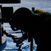 A silhouetted figure is working with what appears to be an ammunition belt or linked rounds on what looks like a naval vessel deck. The person is leaning over a weapon system or mechanical equipment, handling a chain of projectiles or cartridges. The background reveals open ocean water and sky, confirming an at-sea maritime environment.