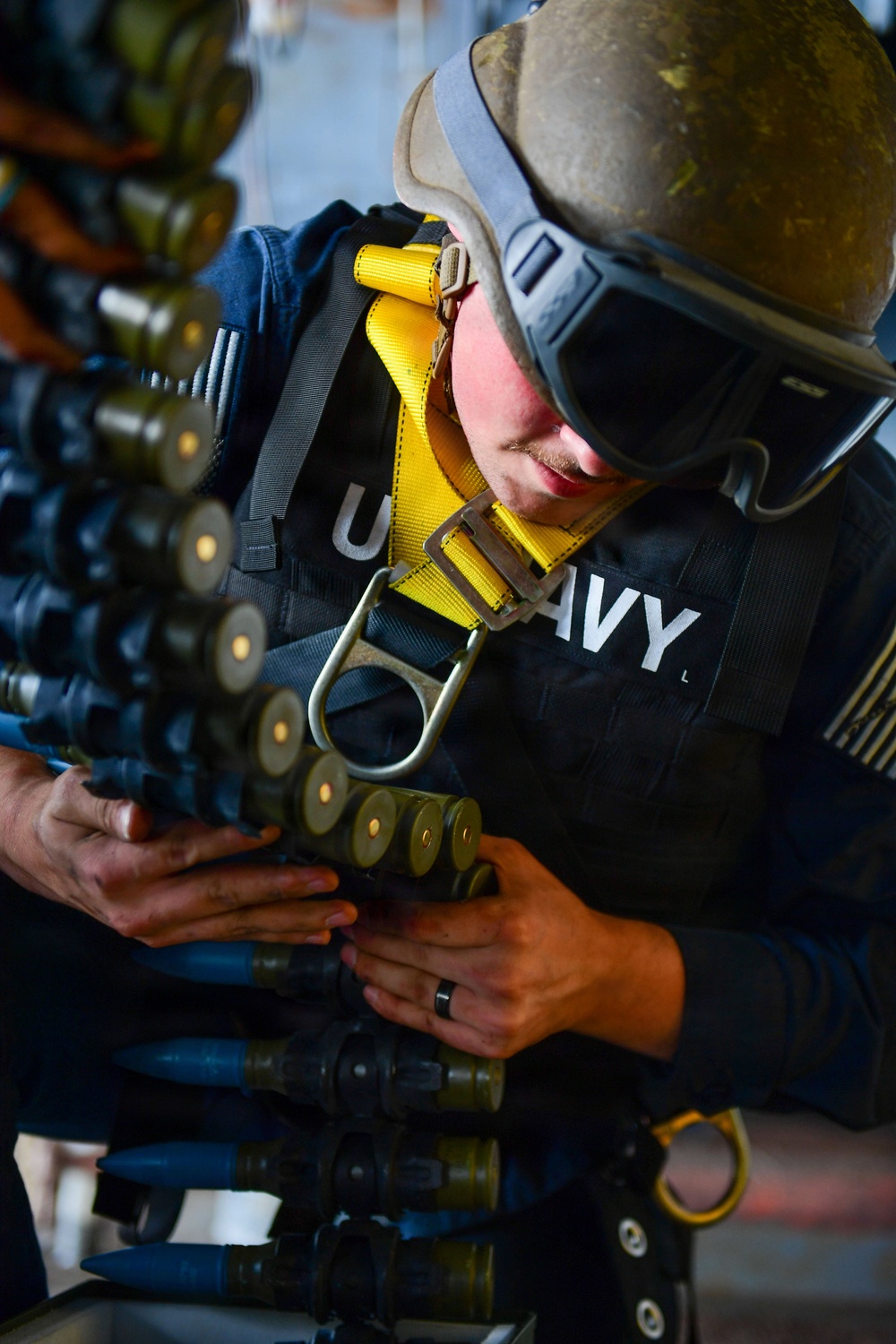 A U.S. Navy sailor wearing a helmet, protective goggles, and a yellow safety harness is handling a linked ammunition belt, likely for a large-caliber weapon system. The sailor appears to be loading or inspecting the ammunition chain, which consists of multiple rounds with visible projectiles and casings. The setting appears to be aboard a naval vessel during an operational or training exercise.