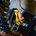 A U.S. Navy sailor wearing a helmet, protective goggles, and a yellow safety harness is handling a linked ammunition belt, likely for a large-caliber weapon system. The sailor appears to be loading or inspecting the ammunition chain, which consists of multiple rounds with visible projectiles and casings. The setting appears to be aboard a naval vessel during an operational or training exercise.