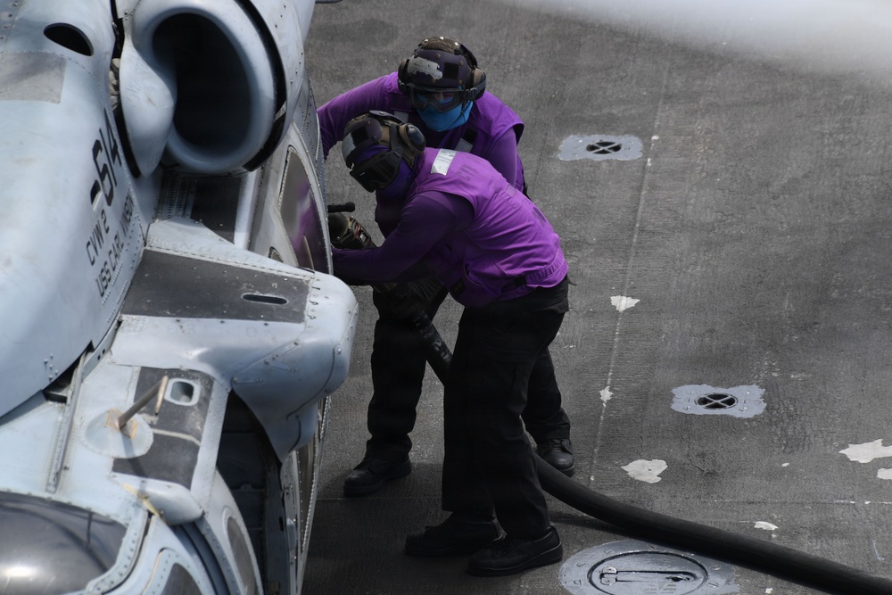 Two naval personnel wearing purple jerseys are fueling a military helicopter on what appears to be an aircraft carrier flight deck. The crew members are handling a fuel hose connected to the aircraft, which bears partial markings including the number '614' and 'USS' text. Purple jerseys in U.S. Navy aviation operations designate aviation fuel handlers (known as 'grapes').
