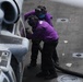 Two naval personnel wearing purple jerseys are fueling a military helicopter on what appears to be an aircraft carrier flight deck. The crew members are handling a fuel hose connected to the aircraft, which bears partial markings including the number '614' and 'USS' text. Purple jerseys in U.S. Navy aviation operations designate aviation fuel handlers (known as 'grapes').