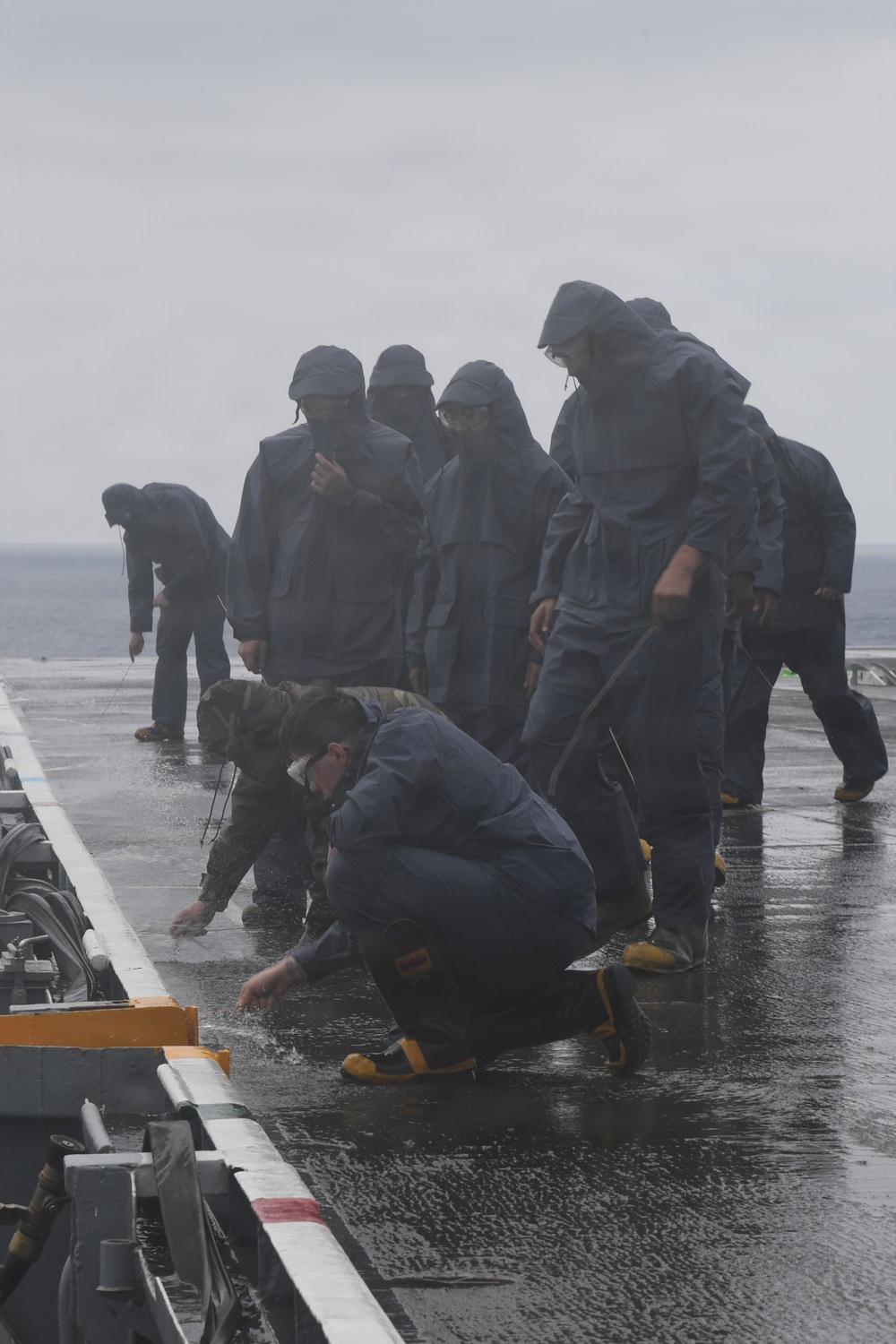 A group of naval personnel in dark blue rain gear and yellow-soled boots are working on a wet flight deck of a warship during rainy, overcast conditions. The sailors appear to be conducting deck cleaning or maintenance operations, with some crouching down and others standing while using tools. The wet, reflective deck surface and open ocean horizon in the background confirm an at-sea operational environment.