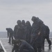 A group of naval personnel in dark blue rain gear and yellow-soled boots are working on a wet flight deck of a warship during rainy, overcast conditions. The sailors appear to be conducting deck cleaning or maintenance operations, with some crouching down and others standing while using tools. The wet, reflective deck surface and open ocean horizon in the background confirm an at-sea operational environment.