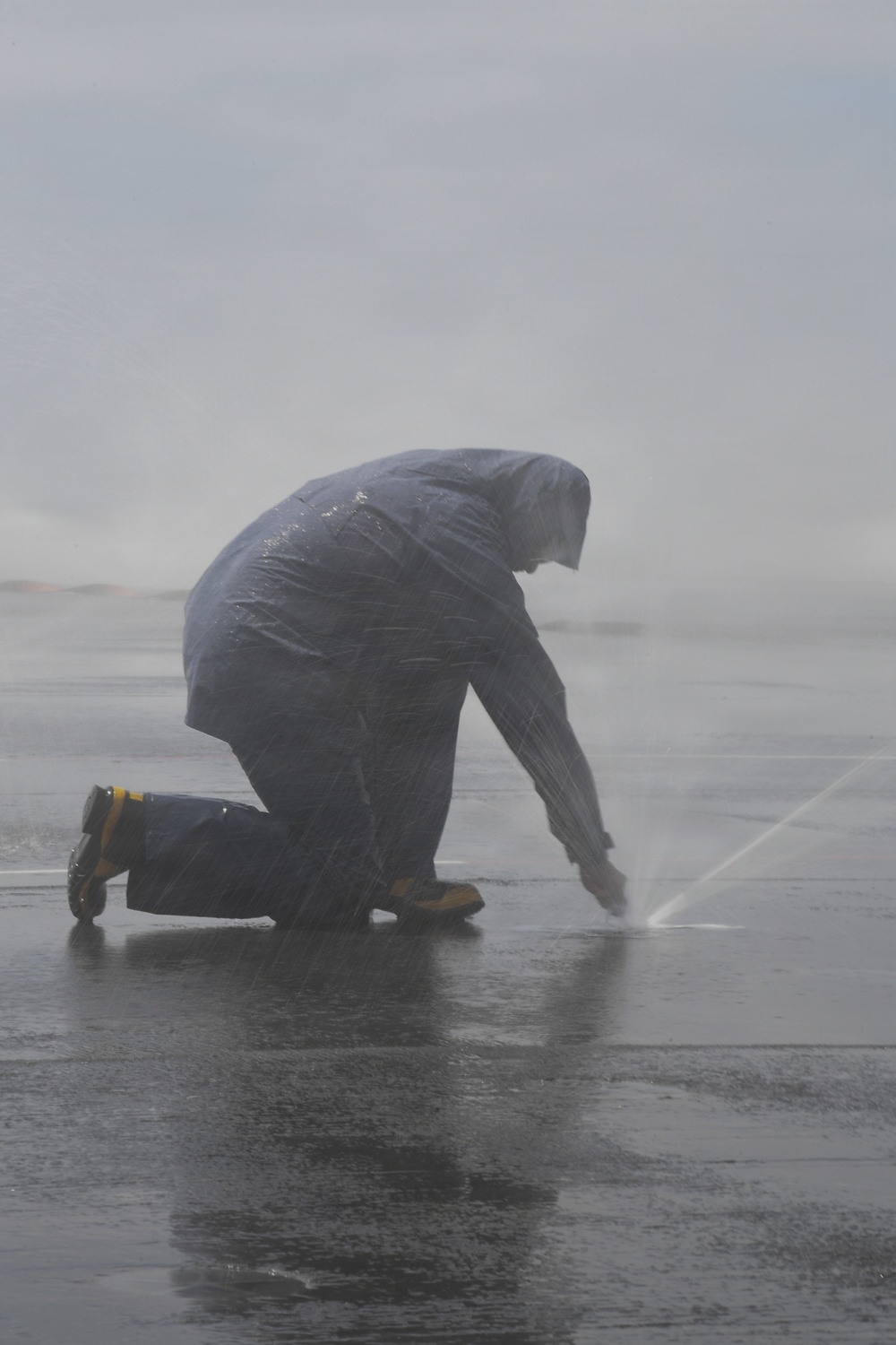 A person wearing a hooded rain jacket and work boots is kneeling on a wet surface in heavy rain or spray conditions. They appear to be working on or near a pressurized water source or leak at ground level, with water jets visibly spraying outward. The scene suggests an outdoor maintenance or emergency response task being performed in harsh weather conditions.