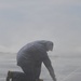 A person wearing a hooded rain jacket and work boots is kneeling on a wet surface in heavy rain or spray conditions. They appear to be working on or near a pressurized water source or leak at ground level, with water jets visibly spraying outward. The scene suggests an outdoor maintenance or emergency response task being performed in harsh weather conditions.