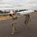 Two military personnel in camouflage uniforms are carrying a fixed-wing unmanned aerial vehicle (UAV) along a paved runway or tarmac. The drone appears to be a small tactical UAV with a white fuselage and orange markings, consistent with military reconnaissance or surveillance platforms. The setting appears to be a rural airfield or training area with dry grassland and tree lines visible in the background.