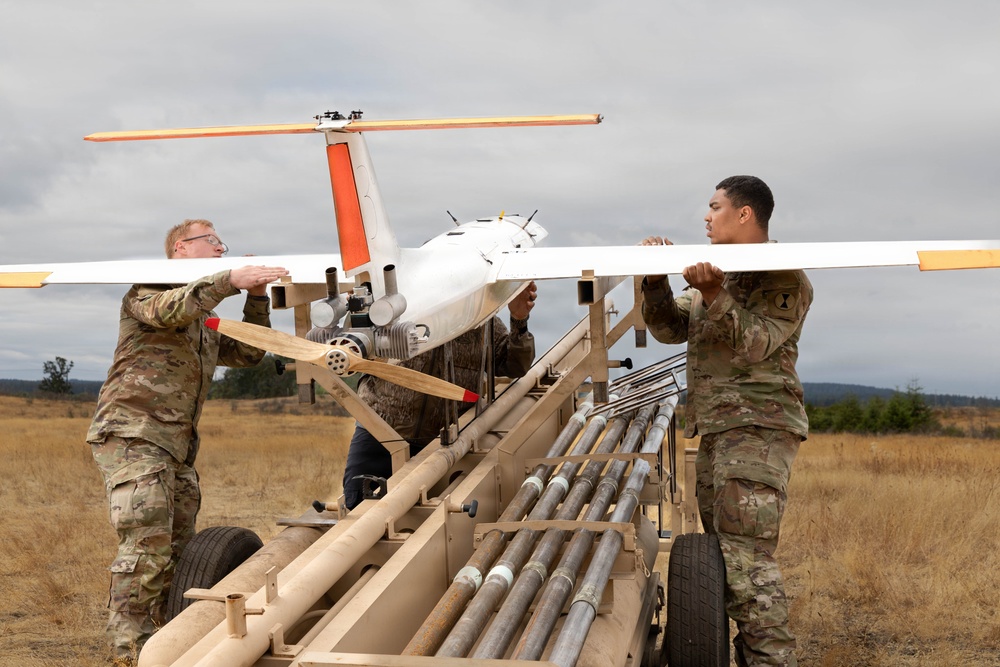 Three military personnel in camouflage uniforms are preparing a fixed-wing unmanned aerial vehicle (UAV) on a pneumatic catapult launch rail system in an open field. The UAV features distinctive orange and white markings with a pusher propeller configuration and appears to be a tactical reconnaissance drone. The setting appears to be a military training or operational area with overcast skies and dry grassland terrain.