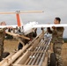 Three military personnel in camouflage uniforms are preparing a fixed-wing unmanned aerial vehicle (UAV) on a pneumatic catapult launch rail system in an open field. The UAV features distinctive orange and white markings with a pusher propeller configuration and appears to be a tactical reconnaissance drone. The setting appears to be a military training or operational area with overcast skies and dry grassland terrain.