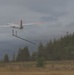A fixed-wing UAV (unmanned aerial vehicle) is captured in flight shortly after launch, with orange-tipped white wings and a rear-mounted pusher propeller configuration. The drone appears to still have a launch rail cradle or catapult bracket attached beneath its fuselage, suggesting it was just launched from the ramp visible in the lower right corner. The setting is an open field surrounded by coniferous trees under an overcast sky.