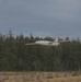 A white fixed-wing aircraft, appearing to be a small UAV or model plane, is captured in low-altitude flight over an open grassy area. The aircraft is flying in front of a dense coniferous forest under an overcast sky. The image has slight motion blur, suggesting the aircraft is moving at speed.