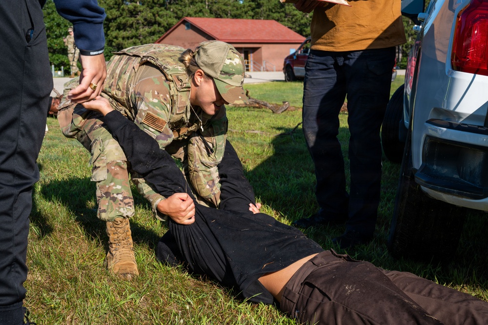 A military or law enforcement training exercise is taking place outdoors on a grassy area. A person in US Army OCP (Operational Camouflage Pattern) uniform is performing a ground control or medical assessment technique on a person lying on the grass. An observer in civilian clothing appears to be taking notes, suggesting this is a supervised training scenario.