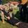 A military or law enforcement training exercise is taking place outdoors on a grassy area. A person in US Army OCP (Operational Camouflage Pattern) uniform is performing a ground control or medical assessment technique on a person lying on the grass. An observer in civilian clothing appears to be taking notes, suggesting this is a supervised training scenario.
