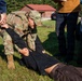 A military or law enforcement training exercise is taking place outdoors on a grassy area. A person in US Army OCP (Operational Camouflage Pattern) uniform is performing a ground control or medical assessment technique on a person lying on the grass. An observer in civilian clothing appears to be taking notes, suggesting this is a supervised training scenario.