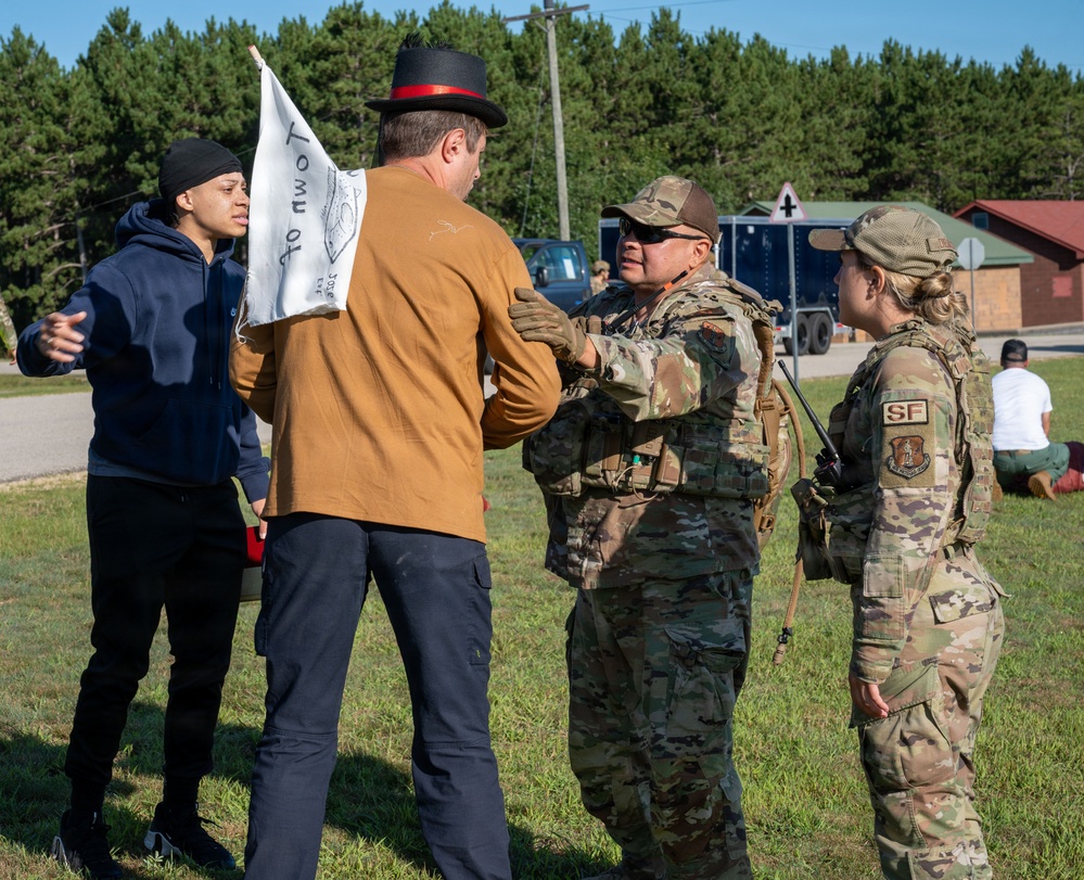 The image shows a military training exercise or security forces scenario involving two U.S. Air Force Security Forces (SF) personnel in OCP camouflage uniforms interacting with two civilians, one of whom is carrying a white sign/banner. The interaction appears to be a simulated crowd control or civil disturbance response exercise, likely conducted at a military installation based on the surrounding infrastructure. The civilians appear to be role-players acting as protesters or demonstrators.