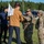 The image shows a military training exercise or security forces scenario involving two U.S. Air Force Security Forces (SF) personnel in OCP camouflage uniforms interacting with two civilians, one of whom is carrying a white sign/banner. The interaction appears to be a simulated crowd control or civil disturbance response exercise, likely conducted at a military installation based on the surrounding infrastructure. The civilians appear to be role-players acting as protesters or demonstrators.