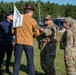 The image shows a military training exercise or security forces scenario involving two U.S. Air Force Security Forces (SF) personnel in OCP camouflage uniforms interacting with two civilians, one of whom is carrying a white sign/banner. The interaction appears to be a simulated crowd control or civil disturbance response exercise, likely conducted at a military installation based on the surrounding infrastructure. The civilians appear to be role-players acting as protesters or demonstrators.