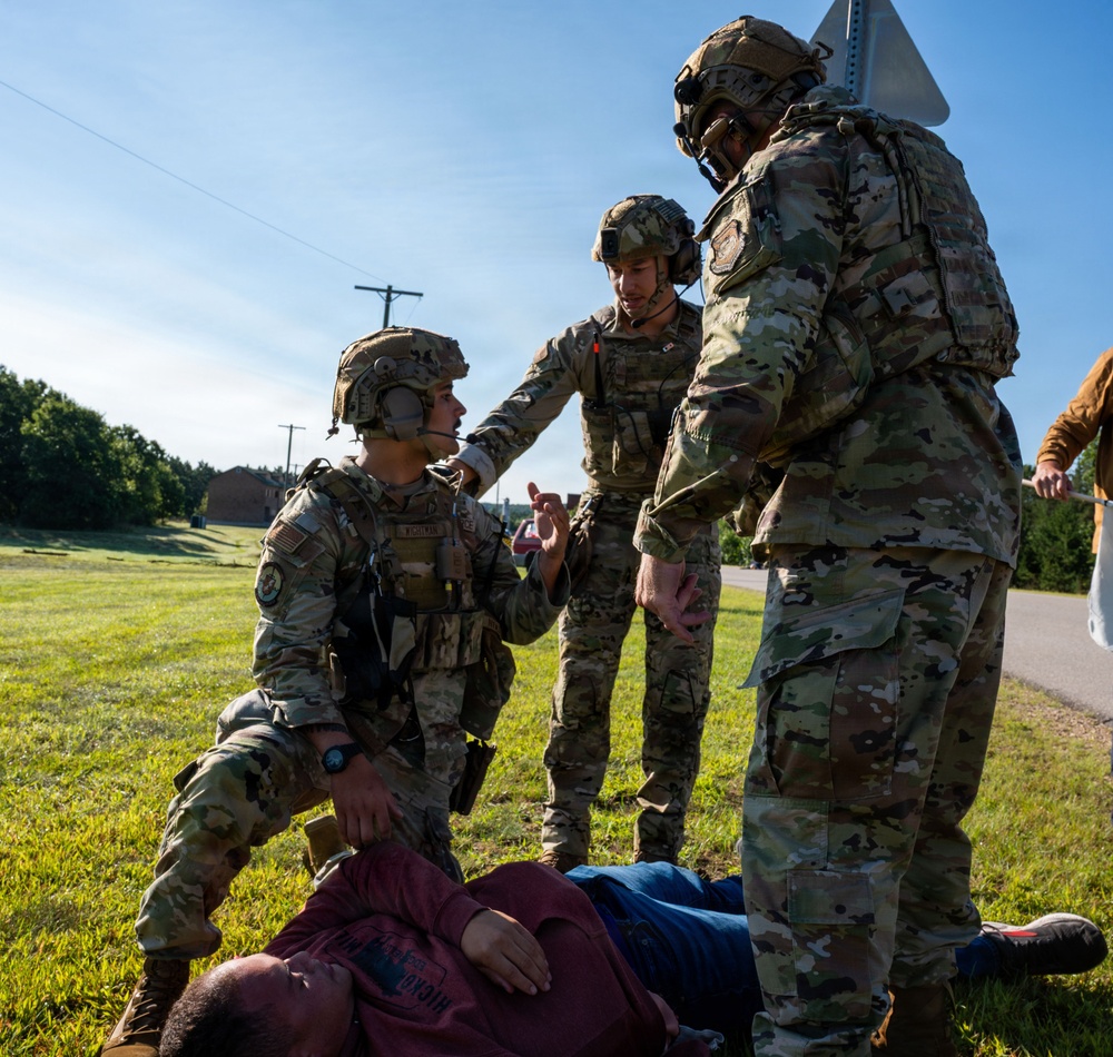 Three military personnel in OCP (Operational Camouflage Pattern) uniforms with tactical helmets and communications gear are attending to a civilian casualty lying on the ground. The scene appears to be a training exercise or simulated rescue/medical response scenario conducted on a grassy field near a road. The soldiers are actively communicating with each other while one kneels beside the downed individual.