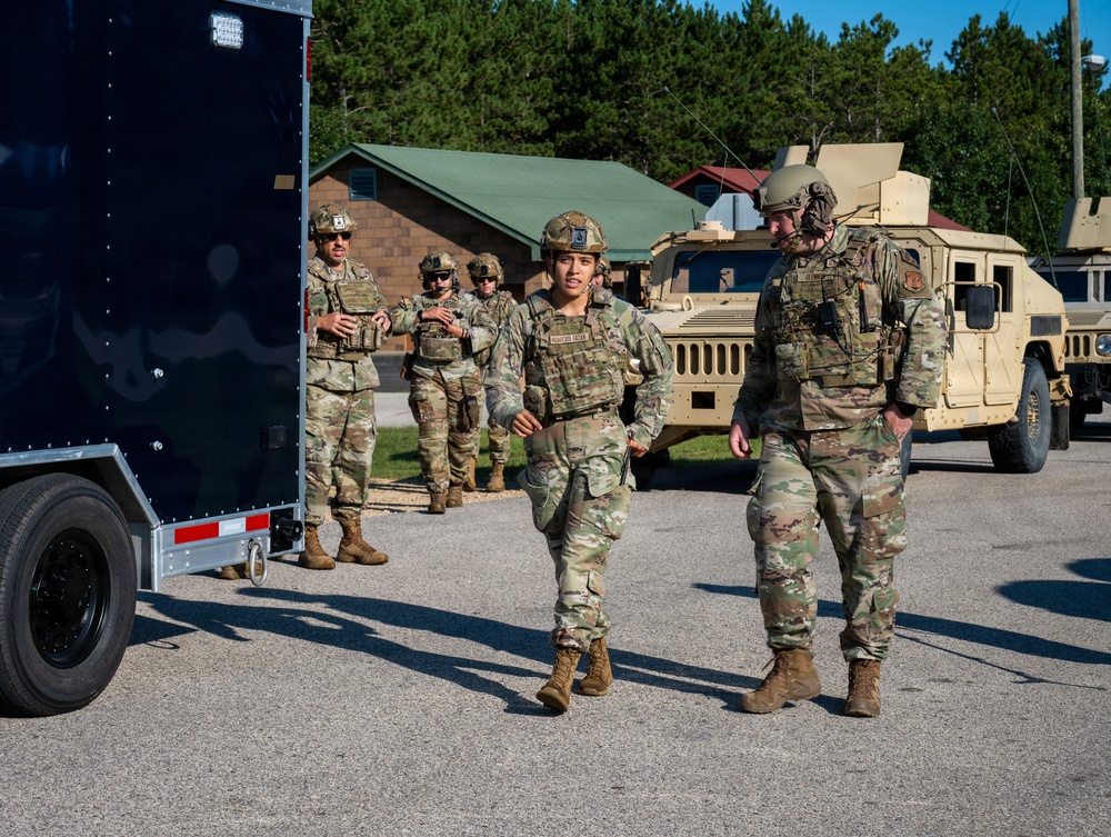 A group of military personnel in full combat gear, including body armor, helmets, and OCP (Operational Camouflage Pattern) uniforms, are gathered in a paved outdoor area near military vehicles. Two soldiers appear to be in active conversation in the foreground while others stand in the background near a dark trailer. The setting appears to be a military installation or training facility with buildings and trees visible.