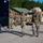 A group of military personnel in full combat gear, including body armor, helmets, and OCP (Operational Camouflage Pattern) uniforms, are gathered in a paved outdoor area near military vehicles. Two soldiers appear to be in active conversation in the foreground while others stand in the background near a dark trailer. The setting appears to be a military installation or training facility with buildings and trees visible.