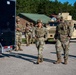 A group of military personnel in full combat gear, including body armor, helmets, and OCP (Operational Camouflage Pattern) uniforms, are gathered in a paved outdoor area near military vehicles. Two soldiers appear to be in active conversation in the foreground while others stand in the background near a dark trailer. The setting appears to be a military installation or training facility with buildings and trees visible.