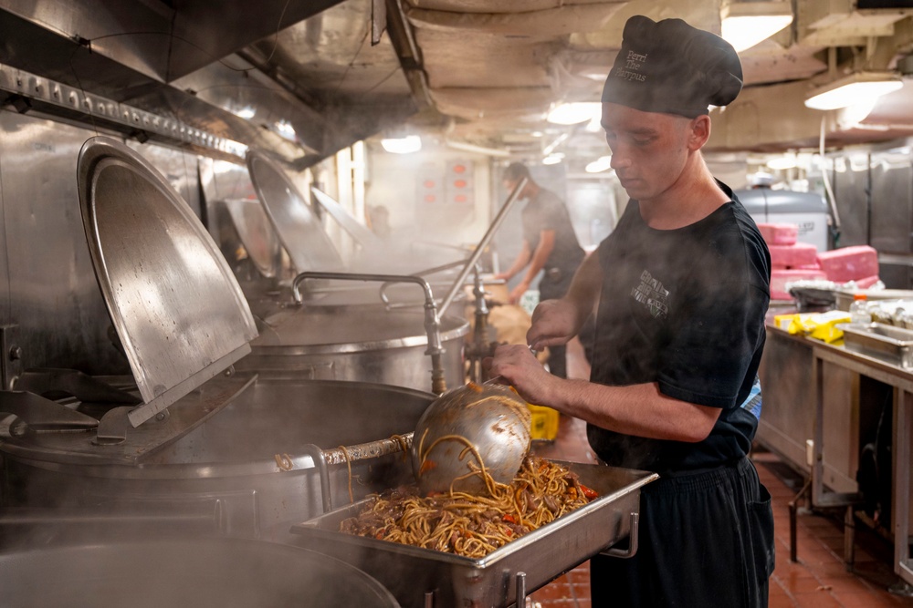 A cook wearing a black chef's hat and dark uniform is working in a large industrial kitchen, ladling noodle dish into a metal serving pan. The kitchen is filled with steam from large cooking vessels, suggesting high-volume food preparation. The setting appears to be a military or institutional galley kitchen based on the heavy-duty stainless steel equipment.