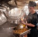 A cook wearing a black chef's hat and dark uniform is working in a large industrial kitchen, ladling noodle dish into a metal serving pan. The kitchen is filled with steam from large cooking vessels, suggesting high-volume food preparation. The setting appears to be a military or institutional galley kitchen based on the heavy-duty stainless steel equipment.