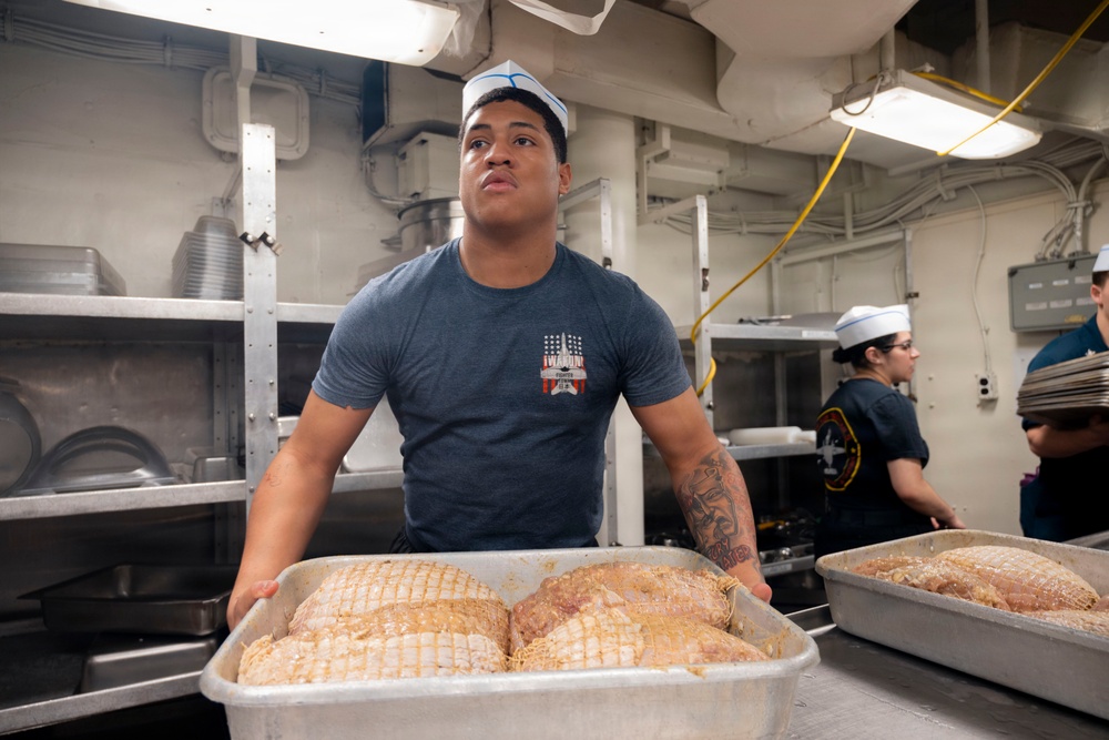 A U.S. Navy sailor wearing a white dixie cup hat and gray t-shirt handles a large metal tray containing multiple netted/trussed turkeys or roasts in what appears to be a military ship's galley kitchen. At least two other personnel in similar uniforms are visible working in the background. The setting suggests holiday meal preparation, likely Thanksgiving, aboard a naval vessel.