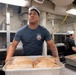 A U.S. Navy sailor wearing a white dixie cup hat and gray t-shirt handles a large metal tray containing multiple netted/trussed turkeys or roasts in what appears to be a military ship's galley kitchen. At least two other personnel in similar uniforms are visible working in the background. The setting suggests holiday meal preparation, likely Thanksgiving, aboard a naval vessel.