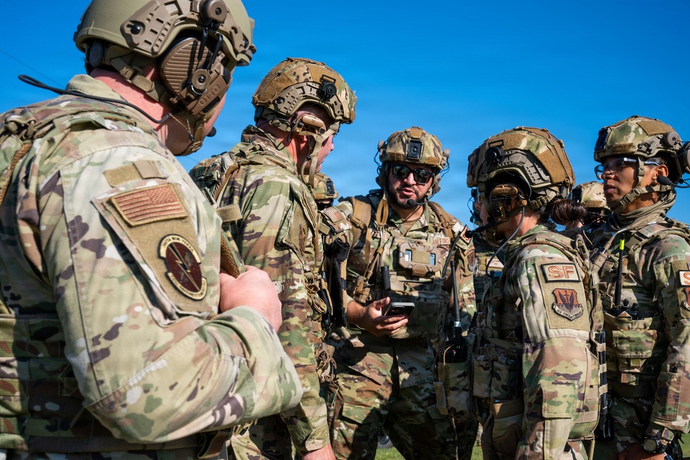 A group of military personnel in full tactical gear are gathered in a briefing or coordination huddle outdoors under a clear blue sky. The soldiers are wearing OCP (Operational Camouflage Pattern) uniforms with tactical helmets, communications equipment, and body armor. Visible patches indicate US Air Force Security Forces (SF) affiliation.
