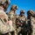 A group of military personnel in full tactical gear are gathered in a briefing or coordination huddle outdoors under a clear blue sky. The soldiers are wearing OCP (Operational Camouflage Pattern) uniforms with tactical helmets, communications equipment, and body armor. Visible patches indicate US Air Force Security Forces (SF) affiliation.