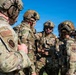A group of military personnel in full tactical gear are gathered in a briefing or coordination huddle outdoors under a clear blue sky. The soldiers are wearing OCP (Operational Camouflage Pattern) uniforms with tactical helmets, communications equipment, and body armor. Visible patches indicate US Air Force Security Forces (SF) affiliation.