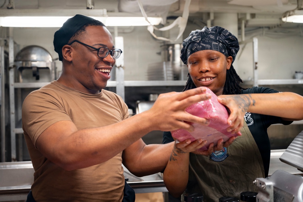Two individuals in a commercial or institutional kitchen are handling a large piece of raw meat wrapped in plastic. Both are smiling and appear to be working together in a food preparation setting. The environment suggests a military galley or large-scale institutional kitchen based on the stainless steel equipment and overhead piping.