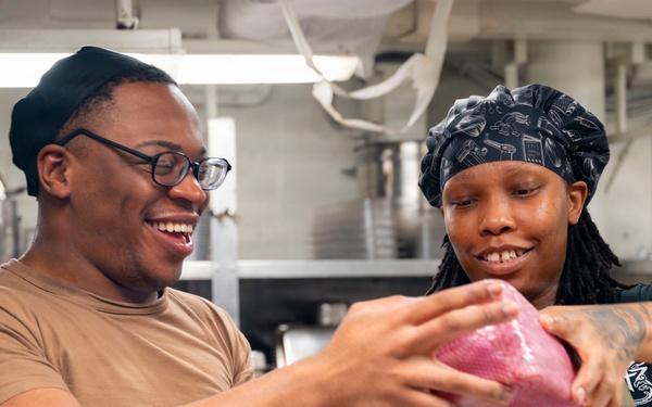 Two individuals in a commercial or institutional kitchen are handling a large piece of raw meat wrapped in plastic. Both are smiling and appear to be working together in a food preparation setting. The environment suggests a military galley or large-scale institutional kitchen based on the stainless steel equipment and overhead piping.
