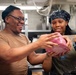 Two individuals in a commercial or institutional kitchen are handling a large piece of raw meat wrapped in plastic. Both are smiling and appear to be working together in a food preparation setting. The environment suggests a military galley or large-scale institutional kitchen based on the stainless steel equipment and overhead piping.
