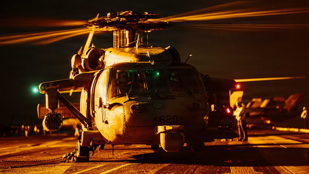A military helicopter, identifiable by the tail number '620', is photographed on what appears to be a naval vessel flight deck during nighttime operations. The scene is dramatically lit with warm orange/amber tones, likely from deck lighting, with rotor blades in motion visible as motion blur streaks. Ground crew personnel are visible in the background attending to flight deck operations.