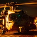 A military helicopter, identifiable by the tail number '620', is photographed on what appears to be a naval vessel flight deck during nighttime operations. The scene is dramatically lit with warm orange/amber tones, likely from deck lighting, with rotor blades in motion visible as motion blur streaks. Ground crew personnel are visible in the background attending to flight deck operations.