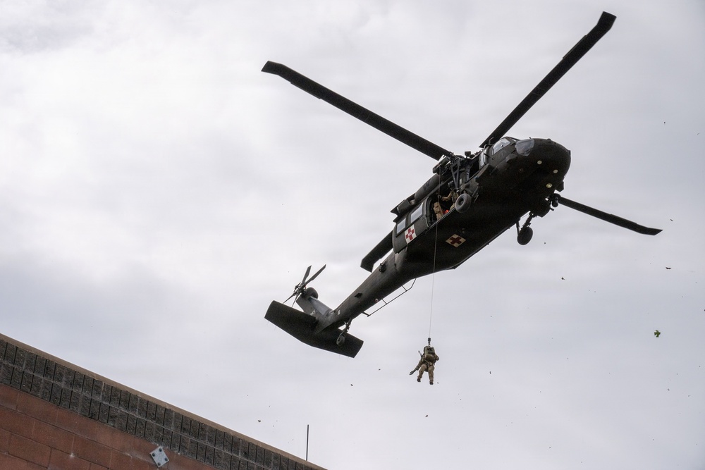A military helicopter, identifiable as a UH-60 Black Hawk with medical cross markings, hovers over what appears to be an urban area. A soldier or personnel is suspended below the aircraft on a hoist or rappel line. The image appears to capture a medical evacuation (MEDEVAC) or rescue training exercise.