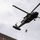 A military helicopter, identifiable as a UH-60 Black Hawk with medical cross markings, hovers over what appears to be an urban area. A soldier or personnel is suspended below the aircraft on a hoist or rappel line. The image appears to capture a medical evacuation (MEDEVAC) or rescue training exercise.