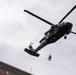 A military helicopter, identifiable as a UH-60 Black Hawk with medical cross markings, hovers over what appears to be an urban area. A soldier or personnel is suspended below the aircraft on a hoist or rappel line. The image appears to capture a medical evacuation (MEDEVAC) or rescue training exercise.