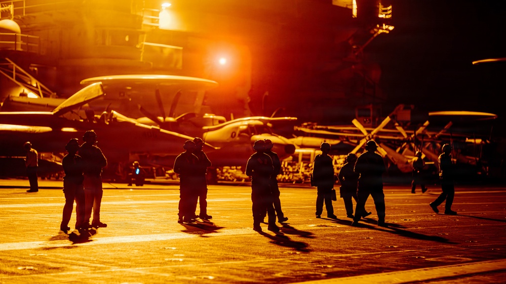 The image shows a naval aircraft carrier flight deck bathed in intense orange-amber light, likely from artificial deck lighting or a setting/rising sun. Multiple silhouetted personnel wearing helmets are visible on the deck alongside parked military aircraft. The dramatic lighting creates strong shadows and a high-contrast silhouette effect across the entire scene.